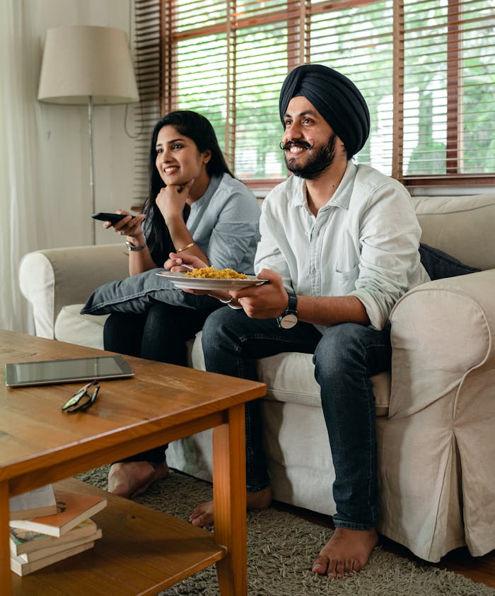 Happy Indian female and bearded male in casual clothes sitting on sofa in living room and smiling while watching tv